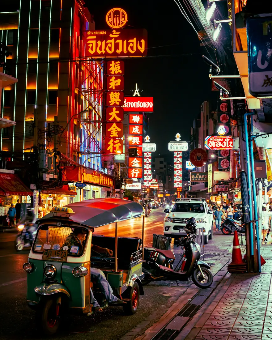 A Bangkok street at night lit by Thai and Chinese neon signs, with a tuk-tuk at the kerb