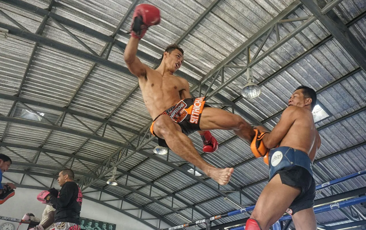 Two Muay Thai fighters mid-bout, one landing a high kick, under a stadium roof