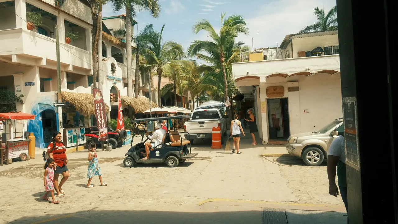 A dusty Punta Mita street with golf carts, parked cars and people walking past low buildings