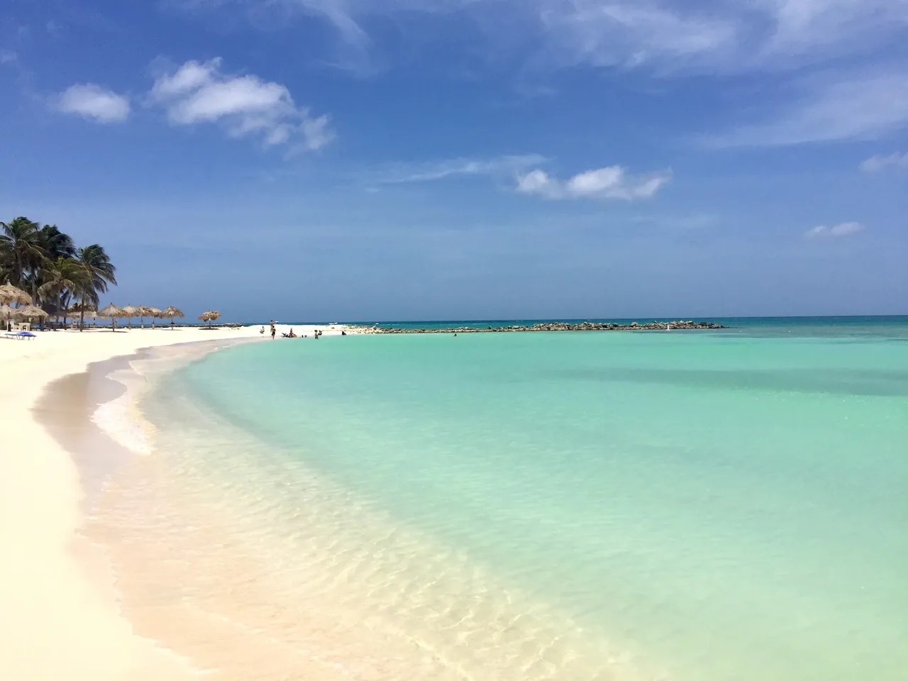 A curving pale-sand beach meeting shallow green water, with palms and low buildings along the shore